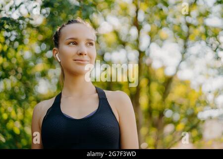 Felice donna dai capelli scuri premurosi in abbigliamento sportivo si guarda via con l'espressione felice si trova nel parco verde dopo il jogging ascolta la musica preferita via Foto Stock