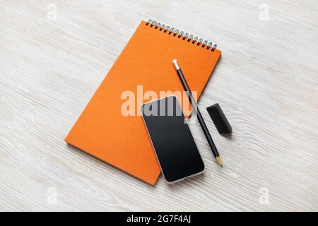 Blocco note arancione vuoto, smartphone, matita e gomma su sfondo tavolo in legno chiaro. Foto Stock