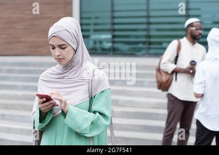 Grave giovane musulmana donna in hijab in piedi contro scala e smartphone di controllo Foto Stock