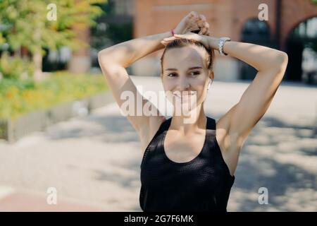 Colpo all'aperto della donna attiva piuttosto sportiva combes coda pony sorride gladfully e guarda a distanza soddisfatto dopo allenamento cardio vestito di nero Foto Stock