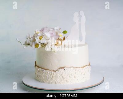 Torta nuziale beige con fiori e figurine della sposa e dello sposo in cima. Torta nuziale su un piano su sfondo bianco Foto Stock