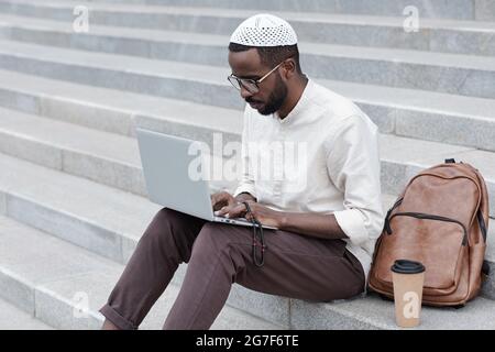 Giovane musulmano nero concentrato in cappellino islamico seduto sulle scale e utilizzando il computer portatile all'aperto Foto Stock