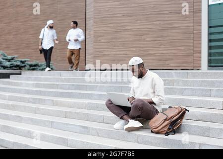 Giovane studente afro-americano concentrato in cappellino kufi seduto con il laptop sulle scale e lavorando al progetto universitario Foto Stock