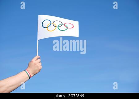 Mano che tiene una bandiera dei Giochi Olimpici fatti a mano all'aperto, contro un cielo limpido, con un bracciale rosso, bianco e verde, colori dell'Ungheria e del Tagikistan na Foto Stock