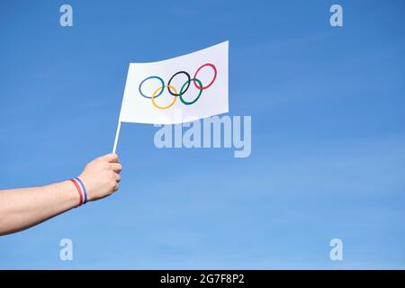 Mano che tiene una bandiera dei Giochi Olimpici fatti a mano all'aperto, contro un cielo limpido, con un bracciale blu, bianco e rosso, colori della bandiera nazionale francese. Foto Stock