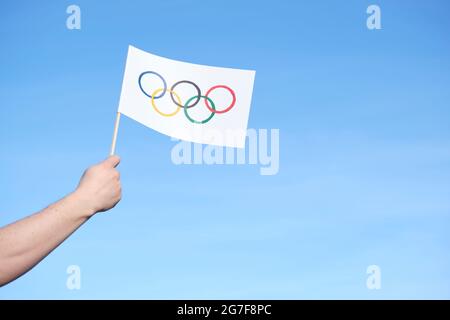 Mano che tiene una bandiera fatta a mano dei Giochi Olimpici all'aperto contro un cielo chiaro in una giornata di sole. Immagine con spazio di copia. Foto Stock