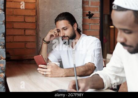 Contenuto giovane ragazzo islamico bearded in camicia bianca seduto al tavolo in caffetteria e scorrendo attraverso i social media sul telefono Foto Stock