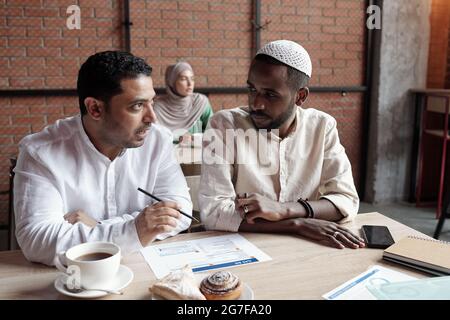 Giovani uomini d'affari multietnici impegnati in abiti islamici seduti al tavolo in un bar e discutere di grafico Foto Stock