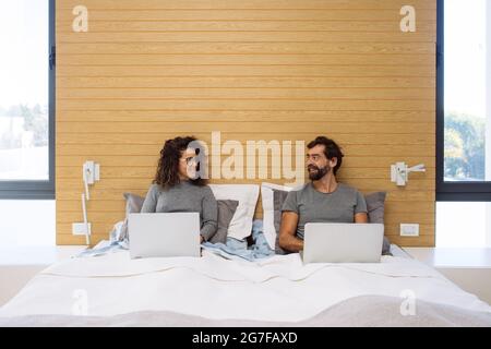 Una coppia in un letto matrimoniale king size, ciascuno con il loro notebook che lavora guardando ciascuno dei loro sreens. Concetto: ufficio domestico che lavora dal letto Foto Stock