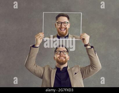 Felice uomo sorridente che tiene una foto di se stesso ridendo isolato su uno sfondo grigio Foto Stock