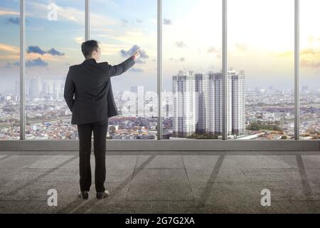 Vista posteriore di un uomo d'affari asiatico che punta attraverso la finestra sulla stanza dell'ufficio Foto Stock