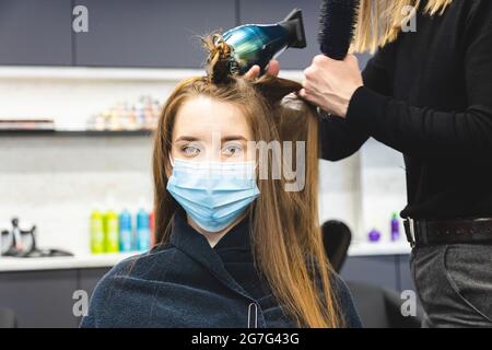 Master donna parrucchiere in una maschera medica asciuga i capelli della ragazza con un asciugacapelli e pettini dopo il lavaggio in un salone di bellezza. Covid-19 pandemia e. Foto Stock