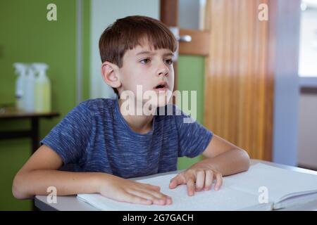 Scolaro caucasico cieco seduto alla scrivania in classe leggendo un libro in braille con le dita Foto Stock