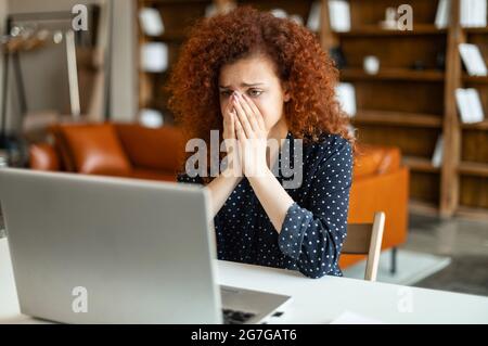 La lotta di scadenza. Giovane lavoratrice da ufficio con capelli ricci stanchi, esausti, e overworked, donna dai capelli rossi seduta con un portatile aperto e chiudendo il viso con le mani Foto Stock