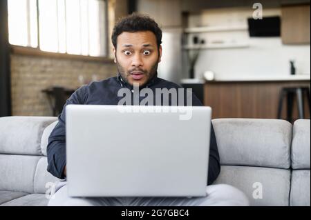 Scioccato giovane indiano che guarda al display del laptop, si sente stupito con una cattiva notizia, licenziato dal lavoro, irritato ragazzo orientale guarda lo schermo del computer e non capisce cosa è successo Foto Stock