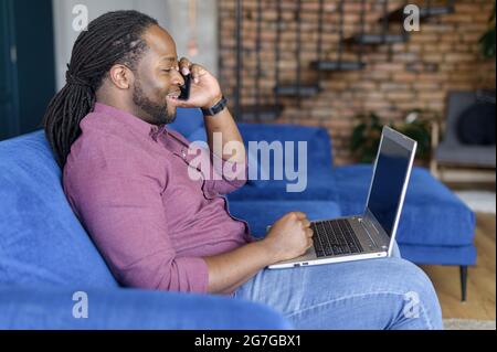 L'allegro afroamericano ha conversazione telefonica, chattando sullo smartphone che lavora con un computer portatile a distanza da casa. Vista laterale Foto Stock