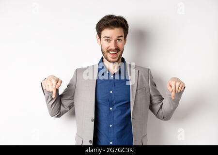 Ehi, dai un'occhiata. Uomo d'affari felice in tuta puntando le dita verso il basso e sorridendo, invitando all'evento, mostrando link, in piedi su sfondo bianco Foto Stock