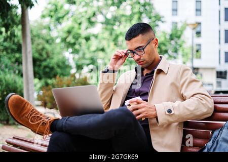 Tutta la lunghezza di giovane e sicuro uomo ispanico in elegante abito casual e occhiali che risponde alla chiamata mentre si siede sulla panca e lavora con il laptop o Foto Stock