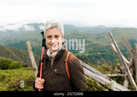 Donna anziana sorridente con zaino e bastone da trekking in piedi sul pendio erboso verso la cima della montagna durante il viaggio in natura guardando la macchina fotografica Foto Stock