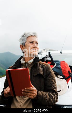 Anziana viaggiatore femminile con capelli corti grigi appoggiati su auto e tablet di navigazione durante il viaggio in campagna e guardando via Foto Stock