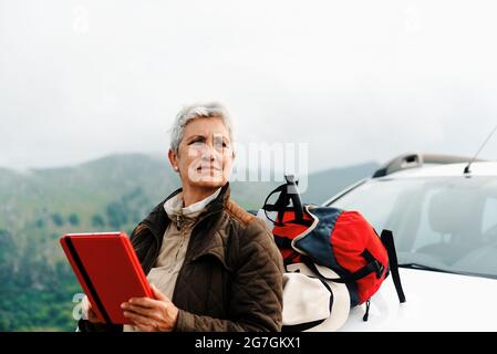 Anziana viaggiatore femminile con capelli corti grigi appoggiati su auto e tablet di navigazione durante il viaggio in campagna e guardando via Foto Stock