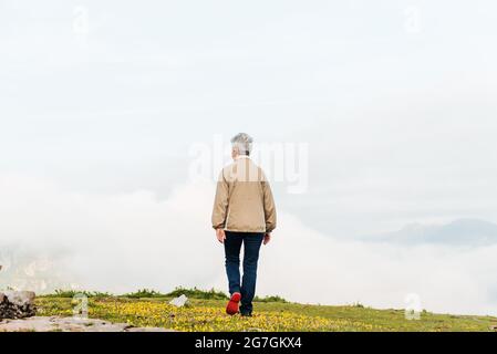 Vista posteriore di una donna anziana irriconoscibile che cammina al mattino su un terreno collinare Foto Stock