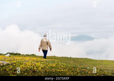 Vista posteriore di una donna anziana irriconoscibile che cammina al mattino su un terreno collinare Foto Stock