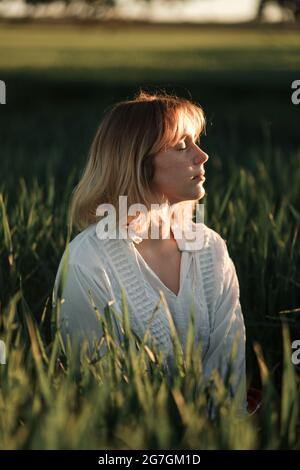 Tranquilla giovane donna in stile retrò blusa bianca seduta tra erba verde alta e occhi chiusi mentre riposa in estate sera in campagna Foto Stock