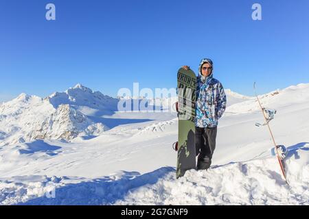 Russia, Elbrus, 16 dicembre 2012: Uno snowboarder con occhiali e un braccio intorno a uno snowboard si trova in cima a una stazione sciistica. Sullo sfondo Foto Stock