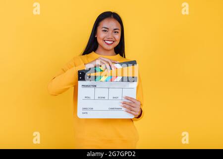 Allegra donna etnica in piedi con clapper film su sfondo giallo in studio e guardando la macchina fotografica Foto Stock