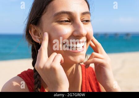 Donna sorridente che applica la lozione del sole sul viso in giornata di sole in estate in spiaggia Foto Stock