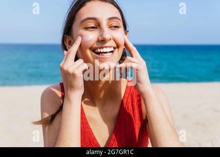 Donna sorridente che applica la lozione del sole sul viso in giornata di sole in estate in spiaggia Foto Stock