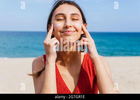 Femmina sorridente con occhi chiusi che applica la lozione del sole sul viso in giornata di sole in estate in spiaggia Foto Stock