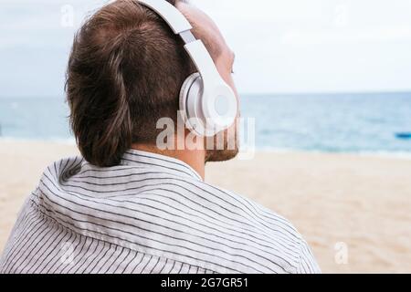 Vista posteriore di un giovane ragazzo spensierato e irriconoscibile in una camicia casual elegante che ascolta la musica tramite le cuffie wireless e goditi una fresca breez Foto Stock