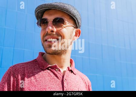 Giovane ragazzo portatore con camicia casual rossa polo e cappuccio guardando la macchina fotografica sopra occhiali da sole e sorridente amichevole mentre si sta in piedi contro parete blu su str Foto Stock