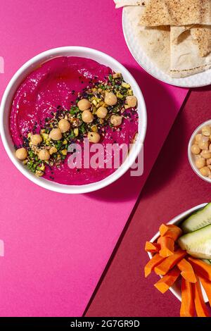 Vista dall'alto dell'appetitoso hummus di barbabietole guarnito con ceci servito su due colori di sfondo con pane e carote fresche e bastoncini di cetrioli Foto Stock
