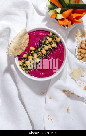 Vista dall'alto dell'appetitoso hummus di barbabietole guarnito con ceci serviti su fondo di tessuto con pane e carote fresche e bastoncini di cetriolo Foto Stock