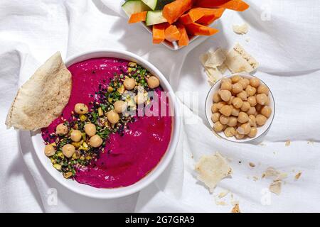 Vista dall'alto dell'appetitoso hummus di barbabietole guarnito con ceci serviti su fondo di tessuto con pane e carote fresche e bastoncini di cetriolo Foto Stock