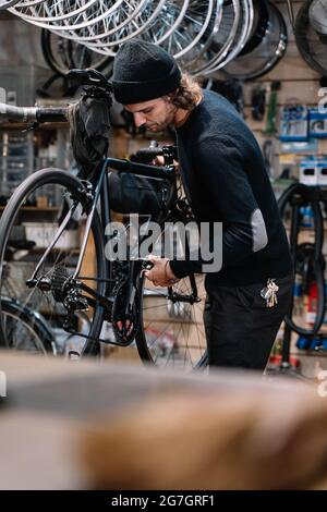 Vista laterale di un giovane meccanico maschio esperto con attrezzo montaggio ruota catena su bicicletta durante gli interventi di riparazione in officina Foto Stock