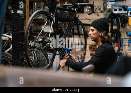 Vista laterale di un giovane meccanico maschio esperto con attrezzo montaggio ruota catena su bicicletta durante gli interventi di riparazione in officina Foto Stock