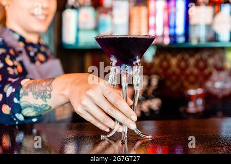 Anonimo sorridente donna barkeeper in piedi al bancone bar con un tipo di bevanda alcolica servita in bicchieri da cocktail creativi a forma di medusa Foto Stock