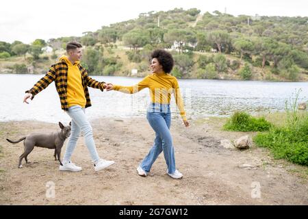 Corpo pieno felice multietnico giovane uomo e donna tenendo le mani e guardando l'un l'altro mentre cammina con il cane sulla costa del fiume in natura Foto Stock