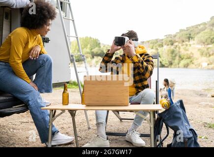 Uomo allegro in occhiali VR che esplora la realtà virtuale vicino a una ragazza nera felice mentre riposa nella natura insieme Foto Stock