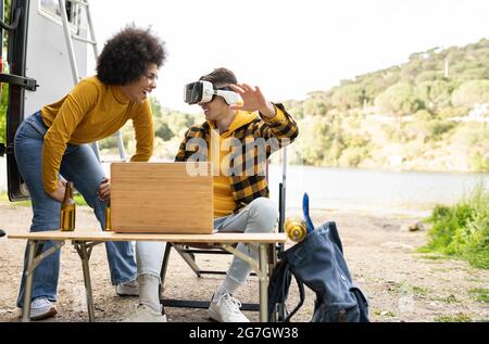 Uomo allegro in occhiali VR che esplora la realtà virtuale vicino a una ragazza nera felice mentre riposa nella natura insieme Foto Stock