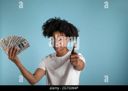 Giovane uomo latino con capelli afro che tengono nuove bollette da cento dollari sorridenti felici e positivi, pollice su facendo eccellente e segno di approvazione Foto Stock