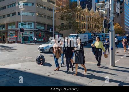 Le persone che attraversano un incrocio di strada nel centro di Sydney, Australia, camminano davanti a una persona inginocchiata e che tiene una tazza che inizia il giorno d'inverno Foto Stock