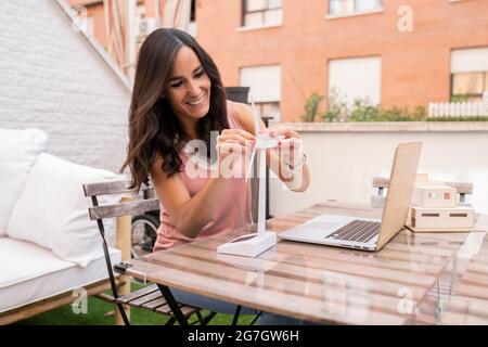 Felice architetto femmina che assembla il modello di plastica del mulino a vento mentre si siede al tavolo con il computer portatile e lavora sul progetto sulla terrazza Foto Stock