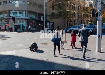 Le persone che attraversano un incrocio di strada nel centro di Sydney, Australia, camminano davanti a una persona inginocchiata e che tiene una tazza che inizia il giorno d'inverno Foto Stock