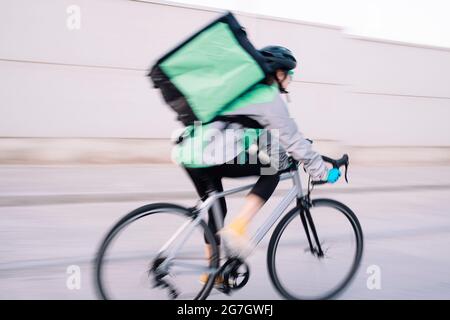 Vista laterale del corriere femminile anonimo con borsa termica in bicicletta sulla strada mentre si consegna il cibo in città, sfocatura movimento Foto Stock