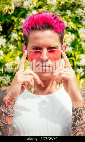 Determinato gay maschile fantasia con capelli rosa tinti e lunghe unghie colorate che toccano occhiali da sole e guardando la macchina fotografica nel parco estivo Foto Stock